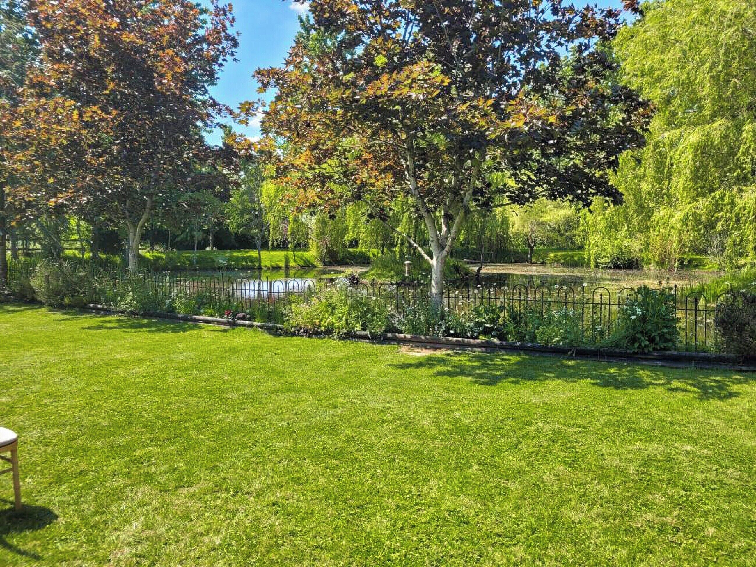 A well-maintained garden with a pond, surrounded by a variety of trees and a metal fence.