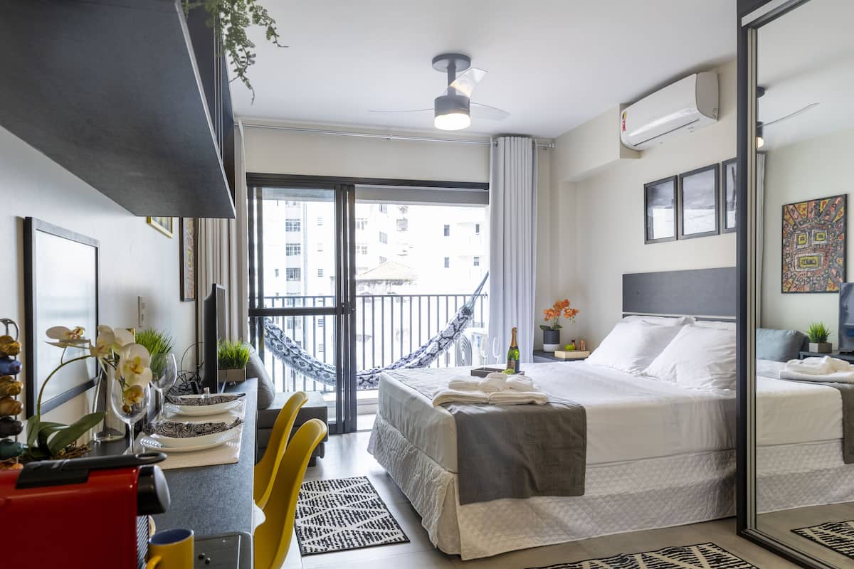 Ein modernes Hotelzimmer mit einem großen Bett, einem Balkon mit Blick auf die Stadt, einem Essbereich mit einem roten Gerät und einem kleinen Tisch mit einer Pflanze.