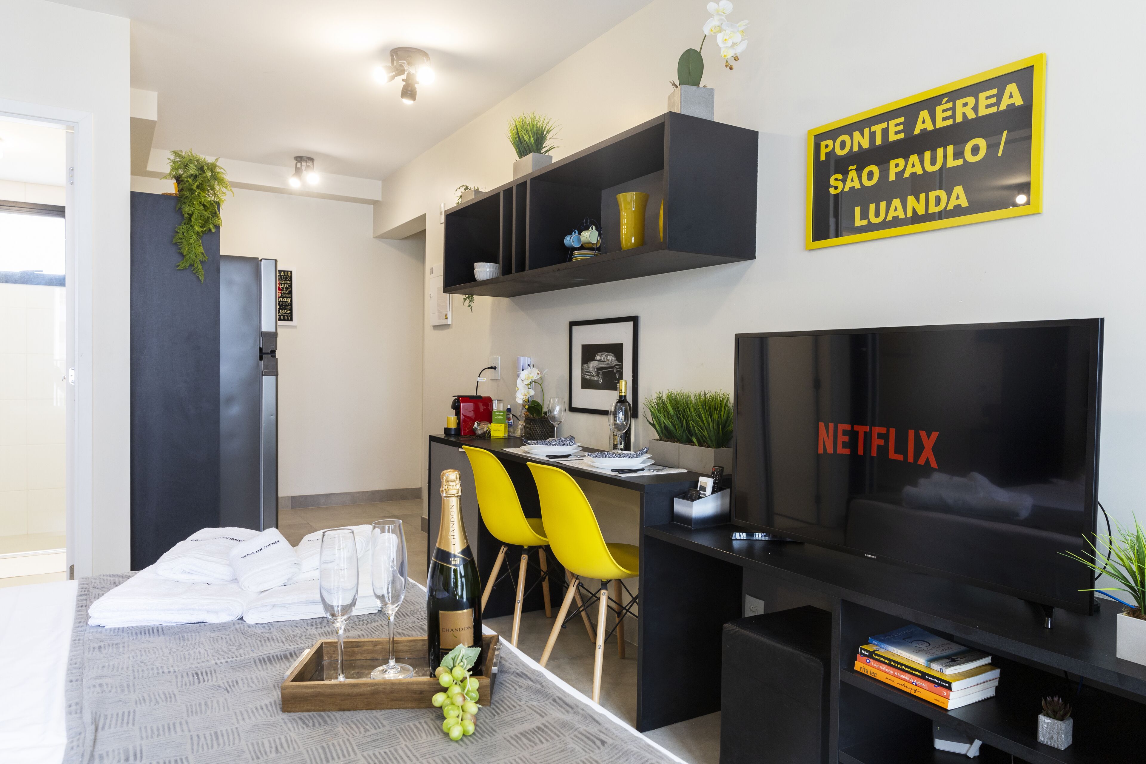 A modern hotel room with a bed, a dining area with a table set for two, a TV, and a sign indicating the hotel is in São Paulo / Luanda.