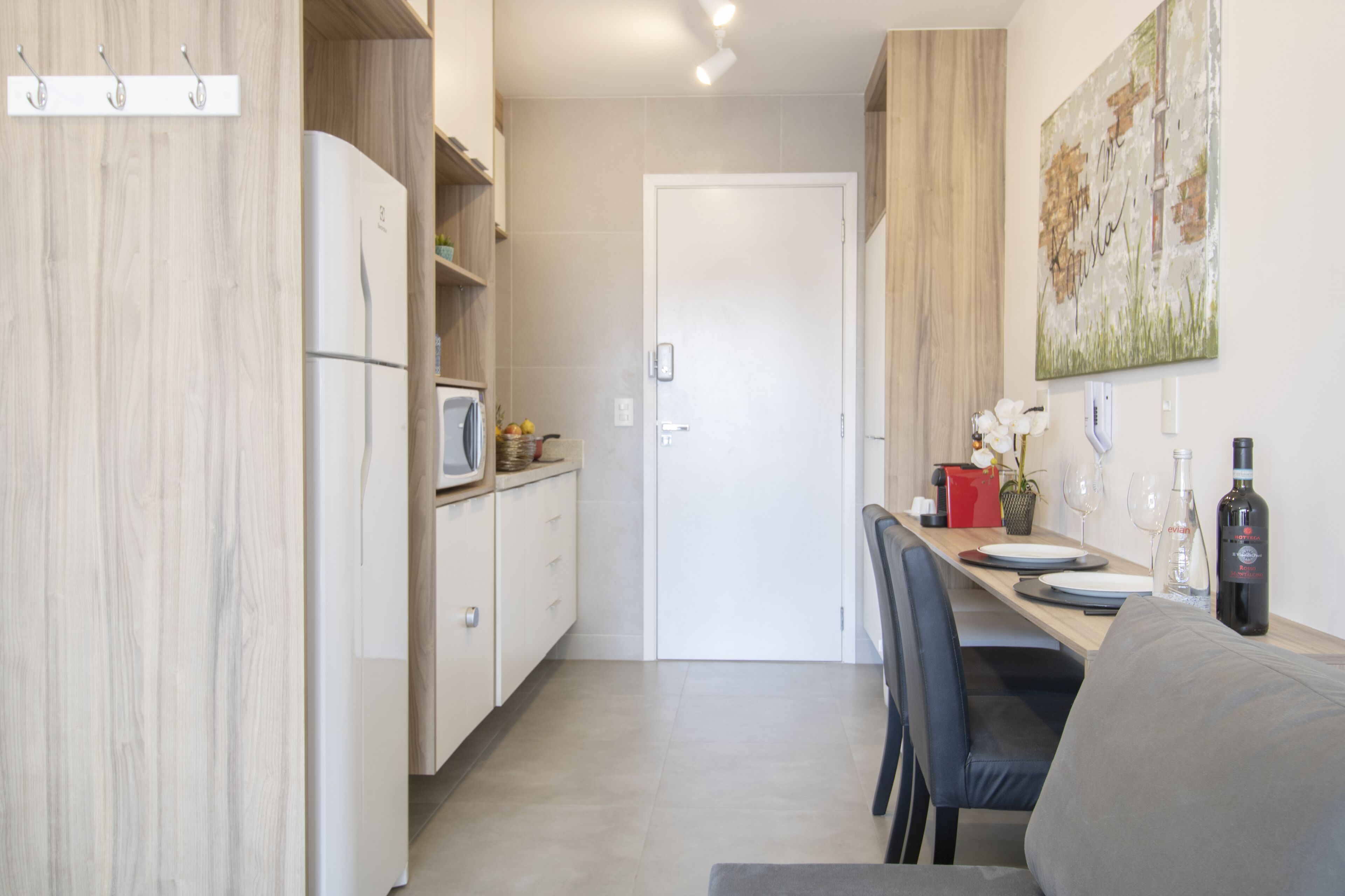 A compact kitchen with a refrigerator, microwave, and wooden cabinets. A dining table with chairs and a small potted plant is visible.