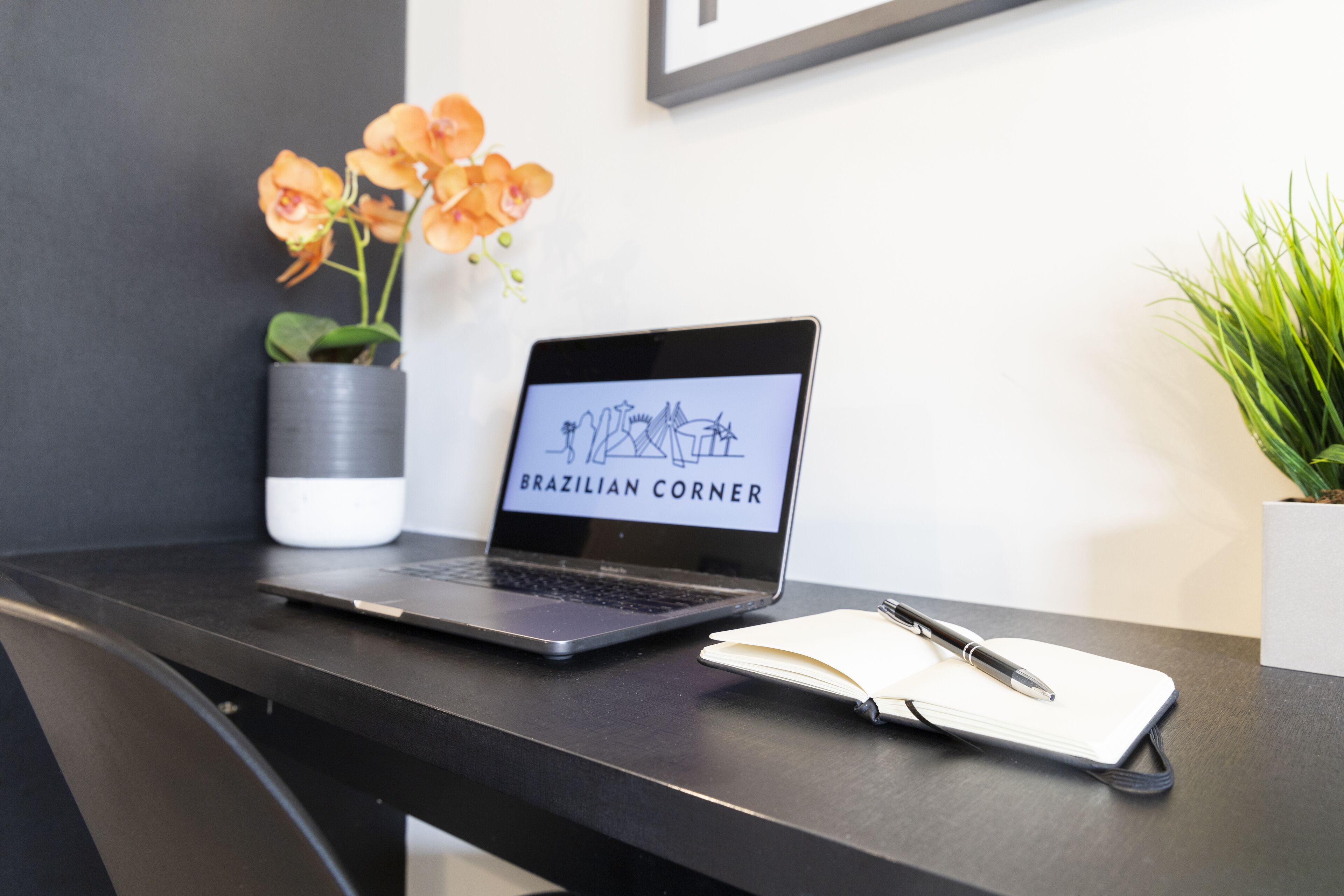 Un ordinateur portable est posé sur un bureau, à côté se trouvent un carnet et un stylo, ainsi qu’une plante en pot et un tableau encadré accroché au mur.