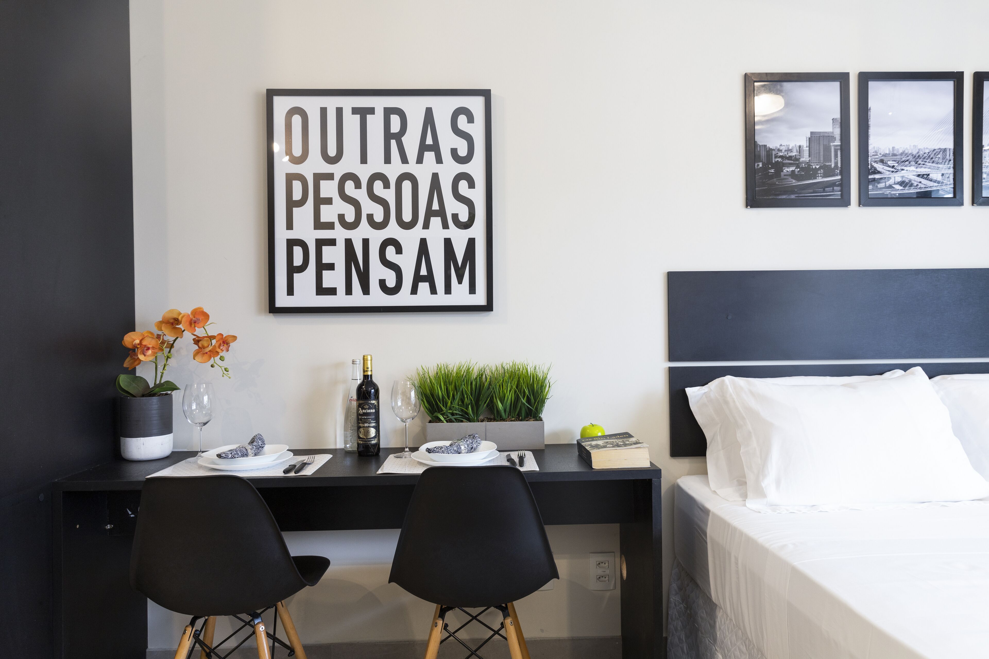 A modern hotel room with a black desk, two black chairs, a bed with white linens, a framed inspirational quote, a small potted plant, a bottle of wine, and a glass.