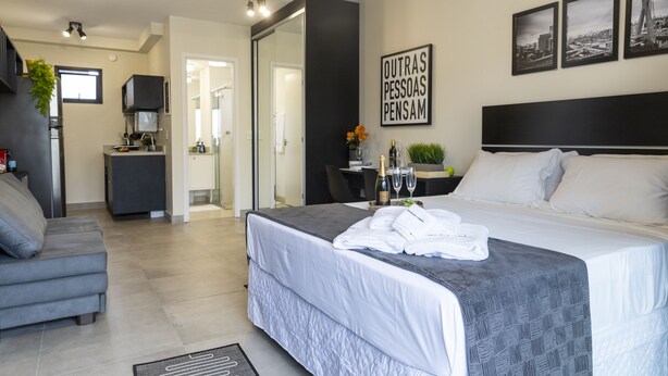 A modern hotel room with a large bed, a grey sofa, a small desk with a chair, and a dining area. - Aurora Paulistana  (São Paulo)