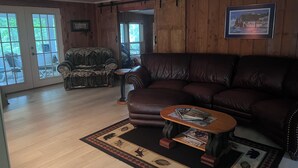 A cozy living room with a brown leather sofa, a patterned armchair, and a coffee table with magazines. A wooden floor and a rug with animal prints are visible. - The Bluebird Cottage.  Just go down 400 feet and jump in lake . (Roscommon)