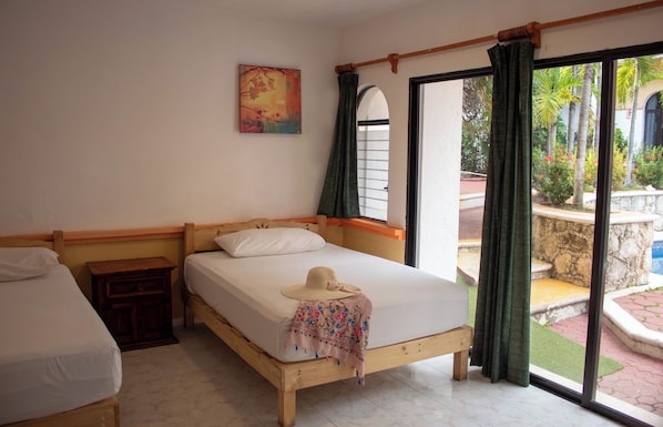 Um quarto com duas camas de madeira, uma janela com cortinas, um quadro na parede e vista para um pátio com plantas e uma estrutura de pedra.
