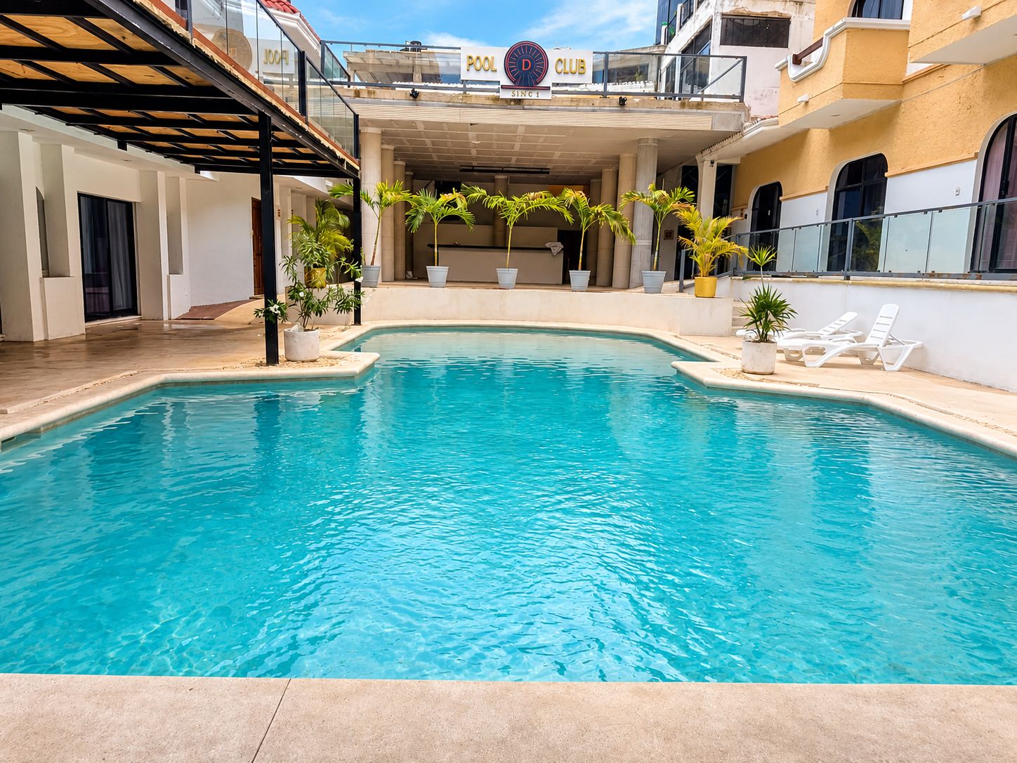An indoor swimming pool with a clear blue water surface, surrounded by potted plants and white lounge chairs.