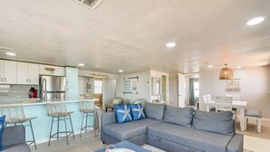 Una sala moderna con un sofá seccional gris, una mesa de centro azul y una zona de cocina con armarios blancos y un refrigerador de acero inoxidable.