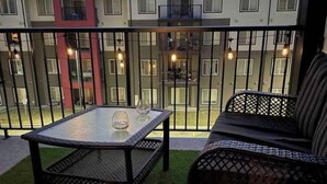 A balcony with a glass table and wicker chairs, overlooking a building with multiple windows and balconies.