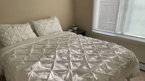 A neatly made bed with a quilted white duvet cover in a room with beige walls and a window with blinds.