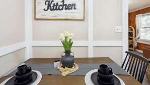 Et spisebord med en vase med hvide blomster, to sorte kopper og et indrammet skilt med teksten 'Kitchen'.