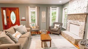 A living room with a stone fireplace, a wooden coffee table, a patterned armchair, and a sofa with decorative pillows. - Mins To AVL, Secluded, Firepit, Family Friendly (Asheville)