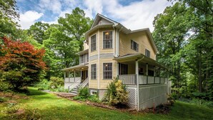 A two-story house with a wrap-around porch, surrounded by lush greenery and trees. - Mins To AVL, Secluded, Firepit, Family Friendly (Asheville)