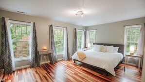 A bedroom with a large bed, multiple windows, a bedside table, and a lamp. - Mins To AVL, Secluded, Firepit, Family Friendly (Asheville)