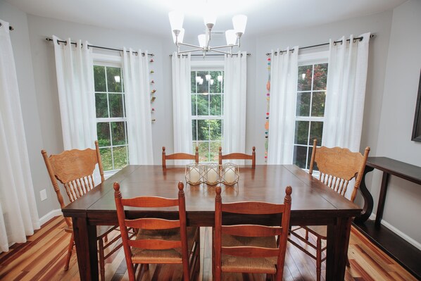 A dining room with a wooden table and chairs, white curtains, and a chandelier.