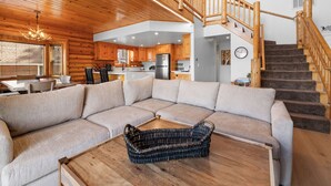 A modern log cabin interior with a large L-shaped sectional sofa, a wooden dining table, and a staircase with wooden railings. - 3 Cubs Lakefront Cabin  (Big Bear Lake)