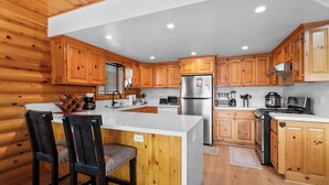 A modern kitchen with wooden cabinetry, a central island with bar stools, stainless steel appliances, and a white countertop. - 3 Cubs Lakefront Cabin  (Big Bear Lake)