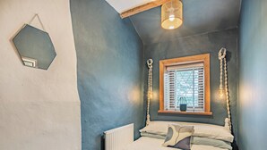 Une chambre avec un plafond en pente, une fenêtre avec des stores, un lit recouvert d’une couverture bleue et un luminaire suspendu.