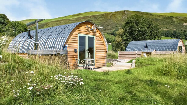 A wooden cabin with a curved roof, a door, and a small patio area with a table and chairs.