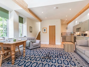 A spacious living area with a wooden dining table, a grey sofa, a patterned rug, and a kitchen in the background. - 1 bedroom accommodation in Dursley (Dursley)