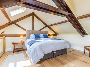 Un dormitorio con techo inclinado, vigas de madera, cama con almohadas, mesita de noche con lámpara y alfombra en el suelo.