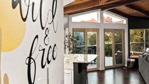 Et moderne interiør med et skilt der siger 'wild & free', træbjælker, en stenvæg og udsigt til en patio med udendørs møbler.