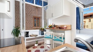 A compact kitchen with a brick wall, white cabinets, a small dining area with a chair, and a view of a balcony with a building. - shinehouse (Sorrento)