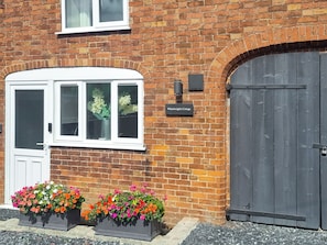 A brick building with a white door and windows, a sign reading "Wheelwright's Cottage", and flower planters in front.