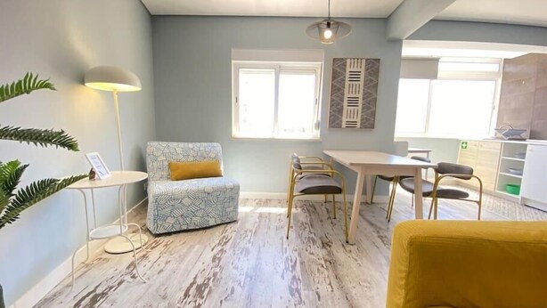 A modern living room with a patterned sofa, a yellow armchair, a dining table, and a kitchen area in the background. - Apartamento Gertrudes (Setúbal)