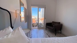 A hotel room with a bed, a chair, a balcony with a view of buildings, and a door. - Apartamento Gertrudes (Setúbal)