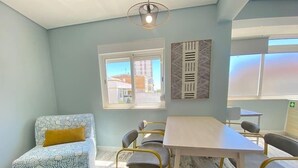 A room with a patterned sofa, a dining table with chairs, a window, and a wall-mounted artwork. - Apartamento Gertrudes (Setúbal)