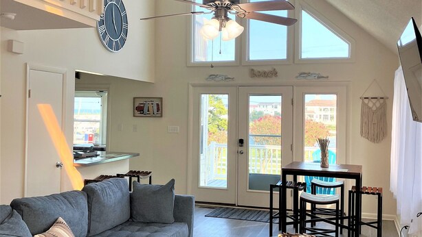 A spacious living room with a gray sectional sofa, a ceiling fan, and large windows allowing natural light. - Surfside Beach Cottage- steps from Virginia Beach Boardwalk! (Virginia Beach)