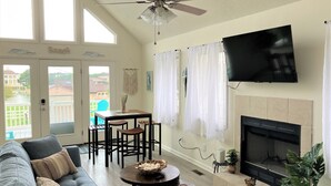 A living room with a gray sofa, a dining table with chairs, a ceiling fan, a TV mounted on the wall, and a fireplace. - Surfside Beach Cottage- steps from Virginia Beach Boardwalk! (Virginia Beach)