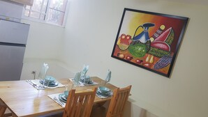 Un comedor con una mesa de madera dispuesta para cuatro personas, un refrigerador y un cuadro de colores en la pared.