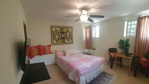 Un dormitorio con una cama, ventilador de techo, televisor montado en la pared, escritorio con silla y ventana con cortinas.