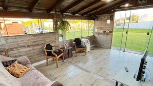 A spacious indoor area with a wooden ceiling, a sofa with a blanket, two chairs, a small table, and a TV on a stand. - Garden house with two bedrooms (Tramandaí Tramandaí)