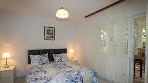 A bedroom with a bed, two bedside lamps, a nightstand, a framed picture on the wall, and a view through a window with curtains. - Green Garden Apartment (Hvar)