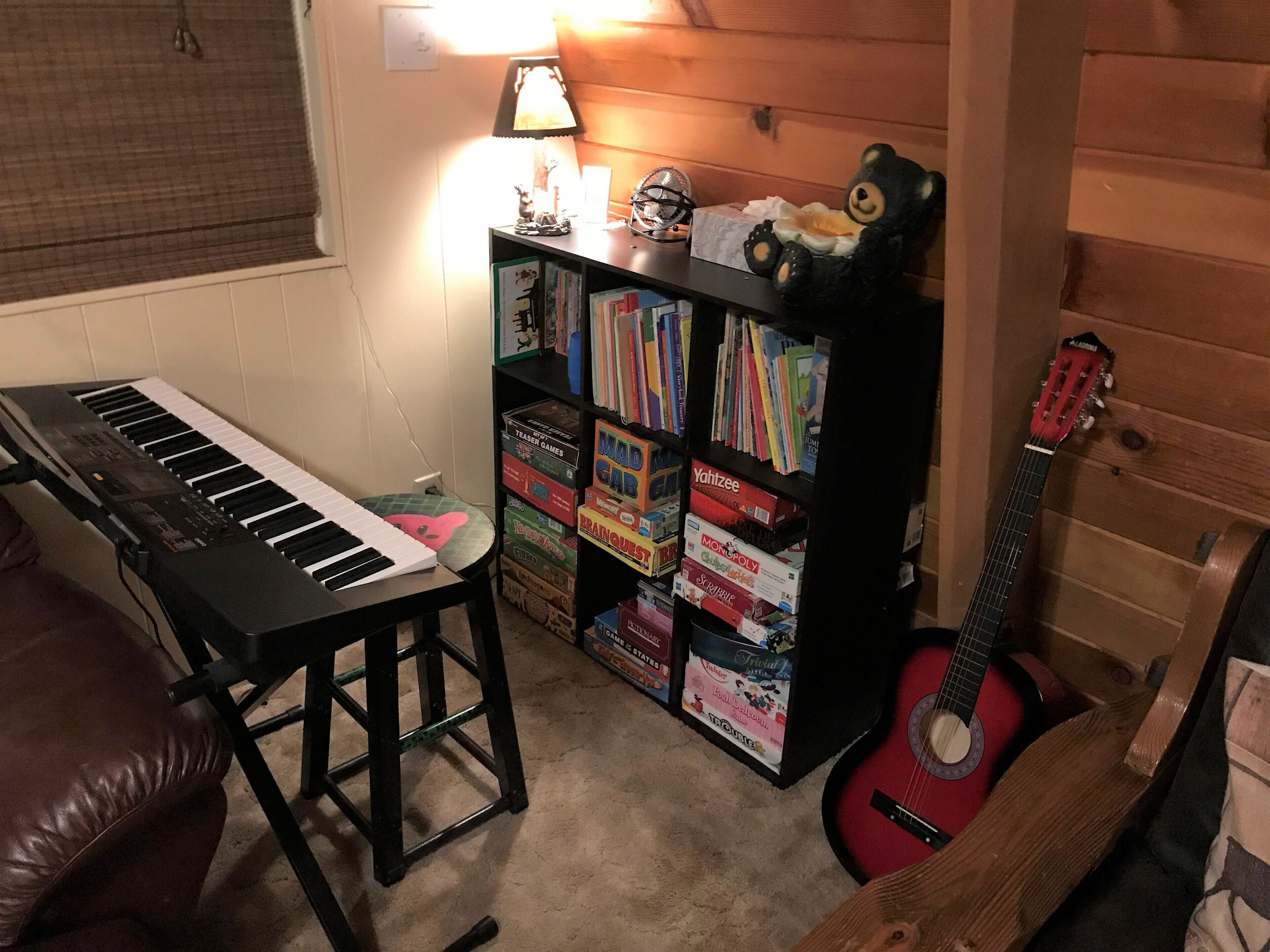 A room with a piano, a bookshelf filled with board games, a guitar, and a teddy bear.