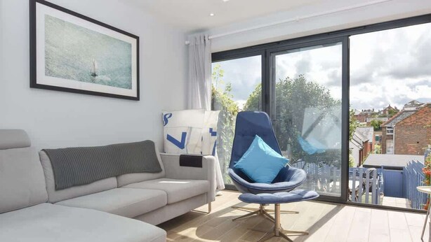 Een moderne woonkamer met een bank, een blauwe stoel met kussen, een groot raam en een schilderij aan de muur.