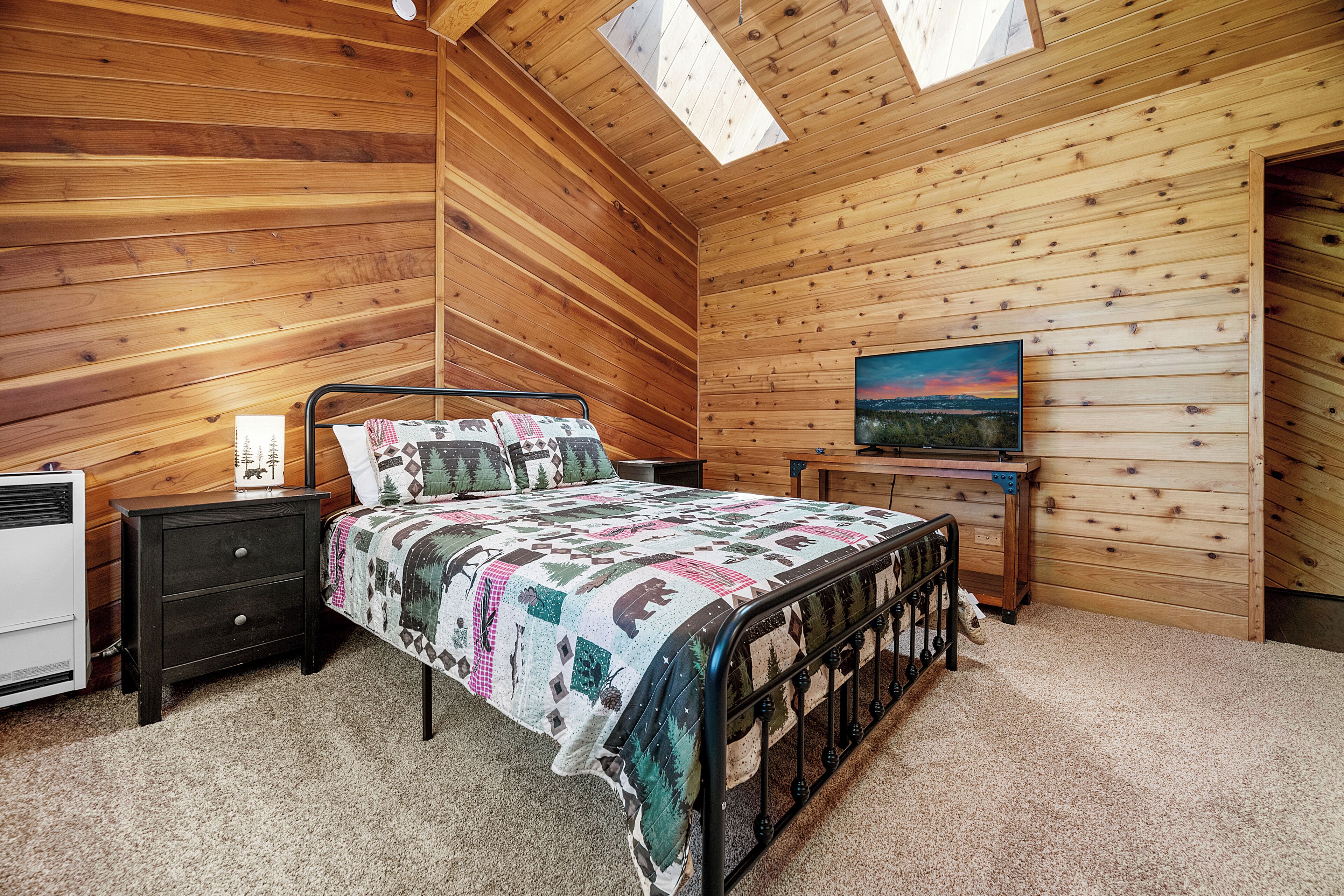 Un dormitorio acogedor con techo inclinado de madera, una cama con colcha estampada, una mesita de noche, una lámpara en la mesita y un cuadro en la pared.
