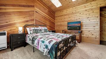 Un dormitorio acogedor con techo inclinado de madera, una cama con colcha estampada, una mesita de noche, una lámpara en la mesita y un cuadro en la pared.