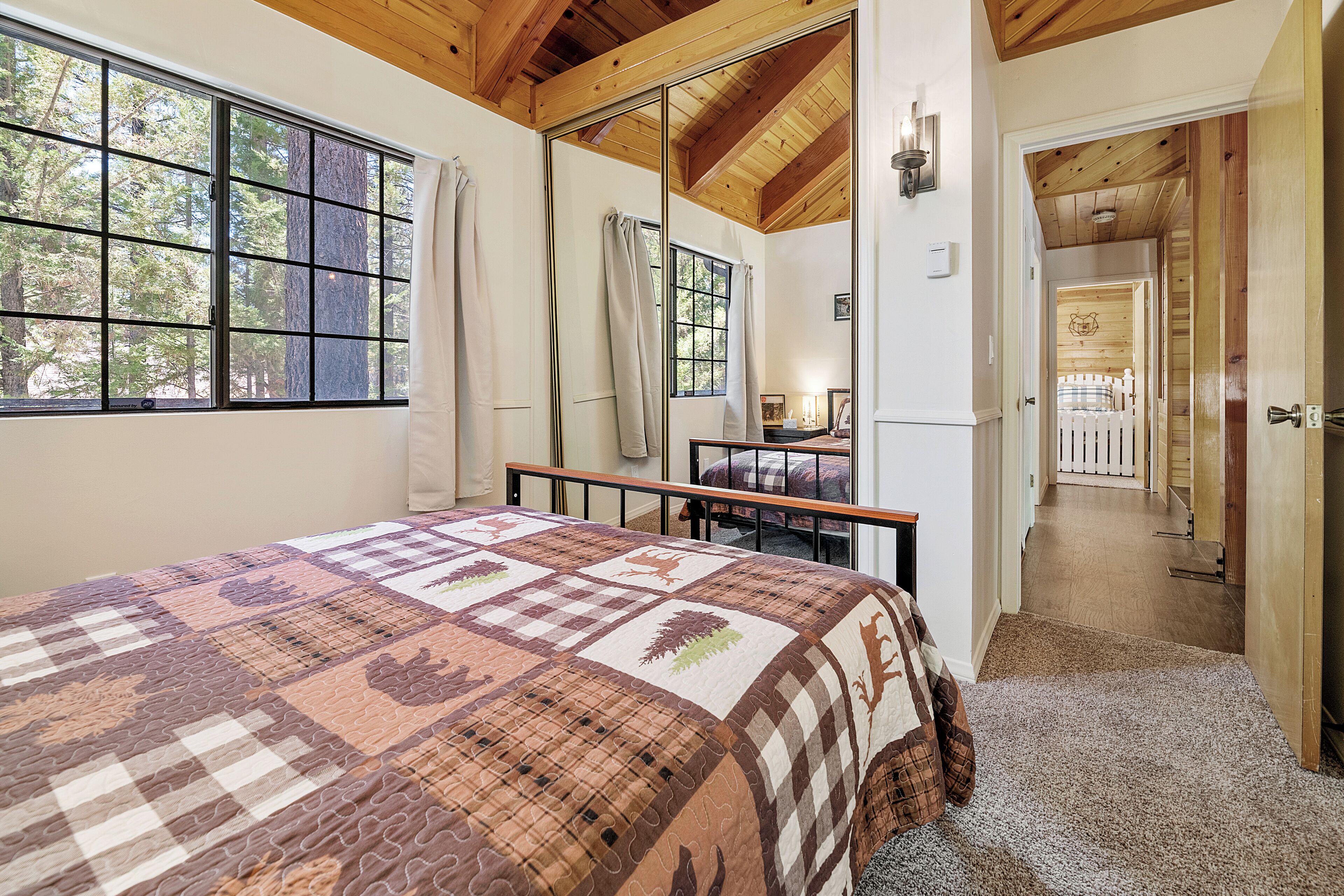 A bedroom with a large bed, a window with curtains, a wooden ceiling, and a view of a deck through an open door.