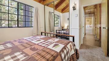 A bedroom with a large bed, a window with curtains, a wooden ceiling, and a view of a deck through an open door.