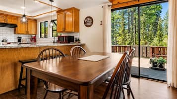 A wooden dining table with chairs, a kitchen with wooden cabinets, a red microwave, and a view of trees through large windows.