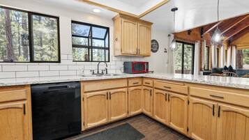 Una cocina con armarios de madera, lavavajillas, microondas y ventanas amplias con vista a un bosque.