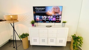Un salon moderne avec un téléviseur à écran plat diffusant un film, un meuble blanc avec des rangements et deux plantes en pot.