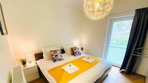 Une chambre d’hôtel moderne avec un grand lit, une table de chevet avec une lampe, une petite plante et une fenêtre donnant sur un paysage verdoyant.