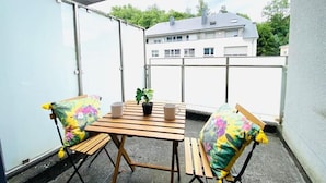 Un balcon avec une table en bois, deux chaises, une plante en pot et une vue sur des bâtiments en arrière-plan.