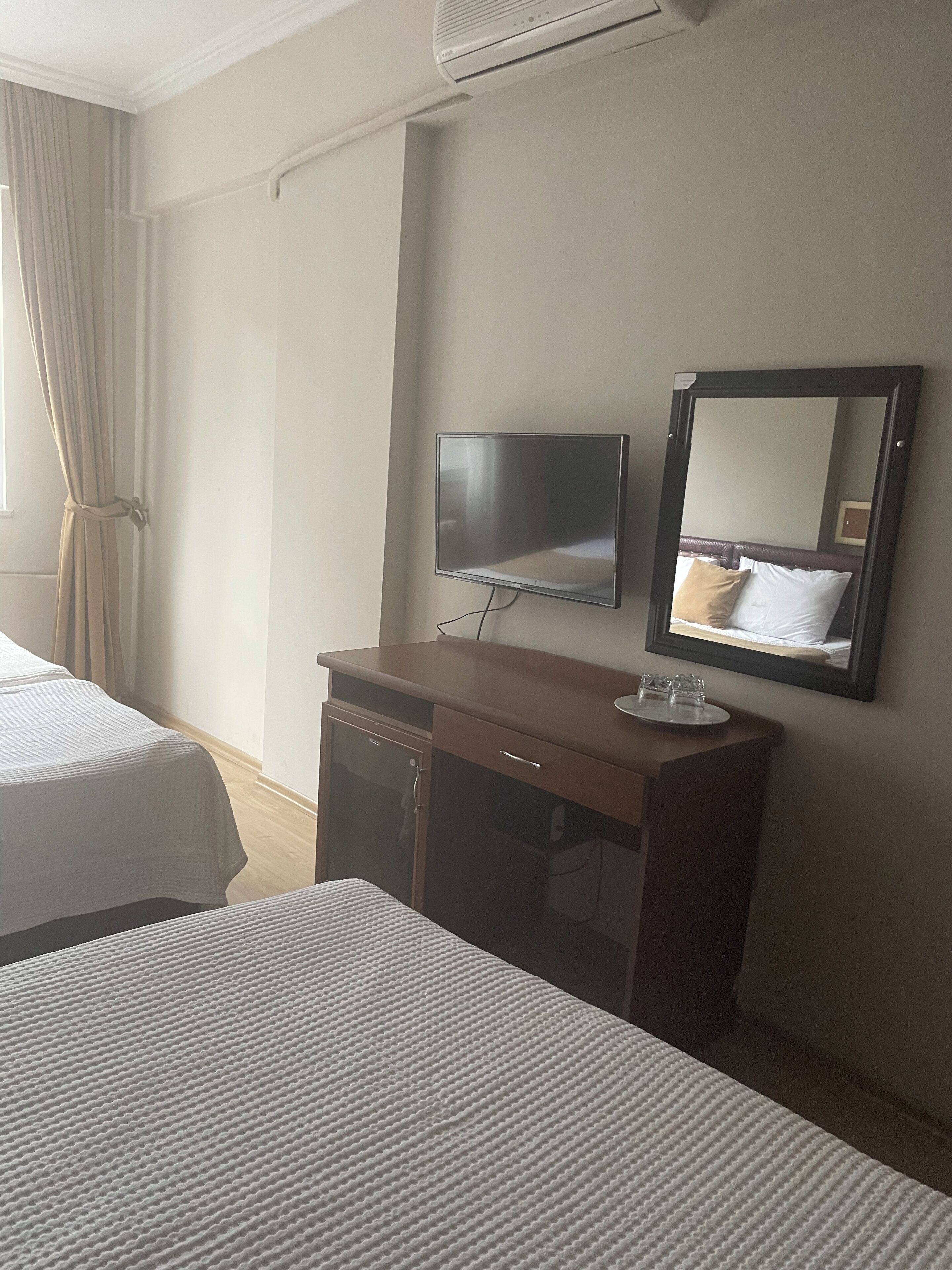 A hotel room with two beds, a desk with a flat-screen TV, a mirror, and a window with curtains.