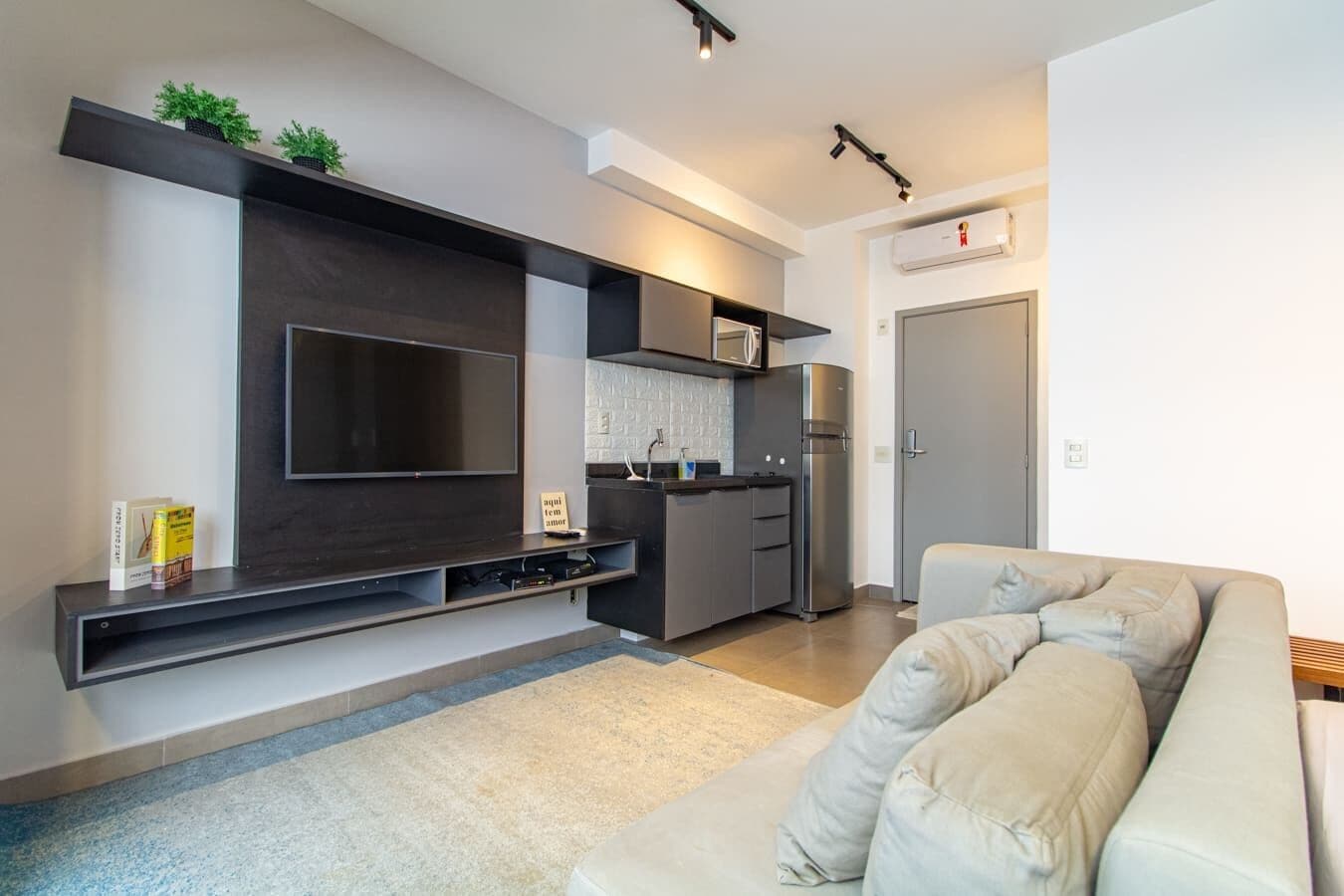 A modern living room with a large flat-screen TV, a built-in kitchen, and a beige sofa.