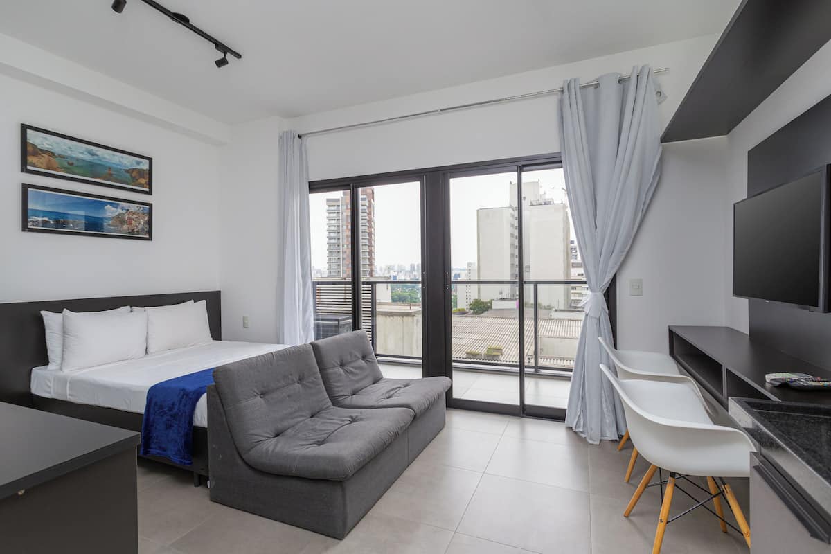 Una habitación de hotel moderna con cama, sofá gris, sillas blancas, televisor y ventanas grandes con cortinas.